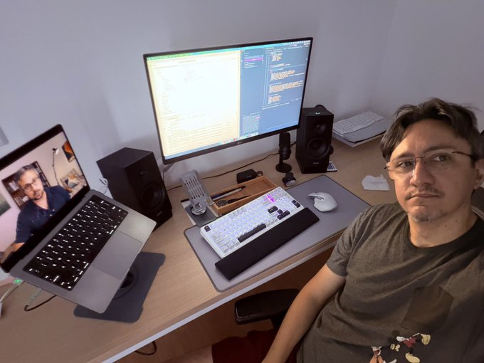 A man with glasses sits at a desk with a large monitor displaying code, a laptop showing a video call, a mechanical keyboard, and studio speakers.