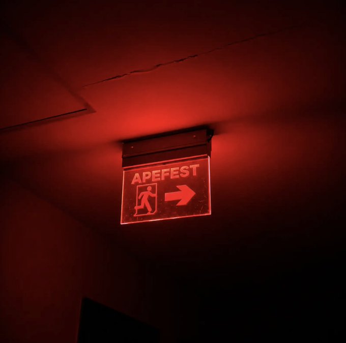 A red illuminated emergency exit sign hangs from a ceiling, displaying the text "APEFEST" alongside a running figure icon and a directional arrow pointing right, tying in perfectly with the post's message that "all signs lead to ApeFest."