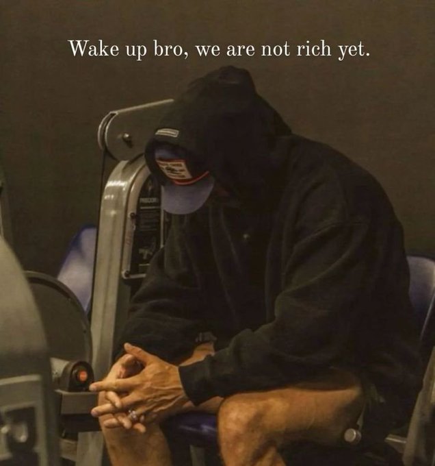 The image features a man sitting down, wearing a black hoodie and hat. He has his hands clasped together while looking at the camera with a sad expression. The caption of the image reads "Wake up bro, we are not rich yet." This phrase is related to the main subject in the image as it seems to be a reference to the idea that one should wake up early and work hard to achieve financial success.