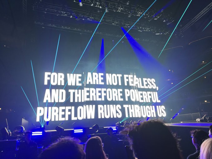 A concert scene with a large illuminated sign reading 'FOR WE ARE NOT FEARLESS, AND THEREFORE POWERFUL PUREFLOW RUNS THROUGH US' bathed in blue laser lights.