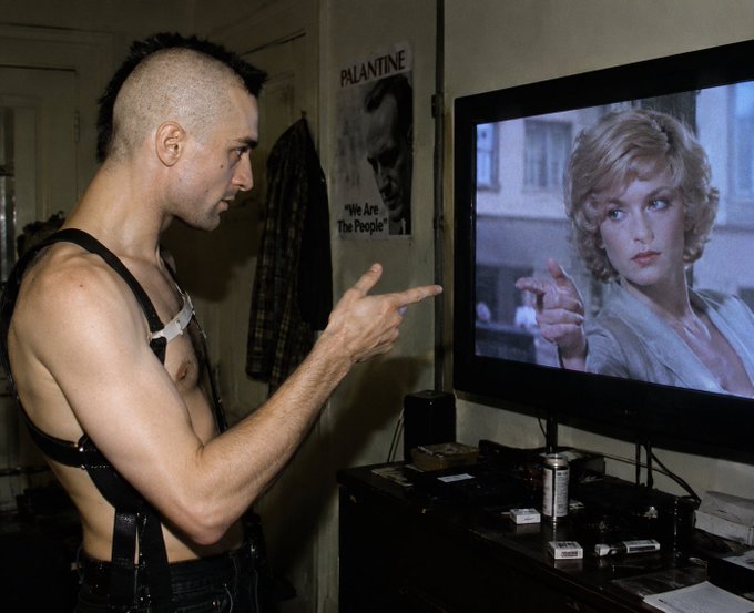 A man with a mohawk wearing a leather harness points at a TV screen displaying a blonde woman, with a 'PALANTINE We Are The People' poster visible on the wall.