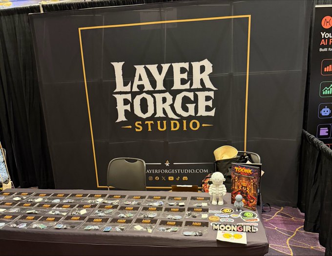 A vendor booth for 'Layer Forge Studio' (layerforgestudio.com) at a convention, displaying artwork, pins, and merchandise including Toonk and Moongirl items, with a small white plush toy on the table.