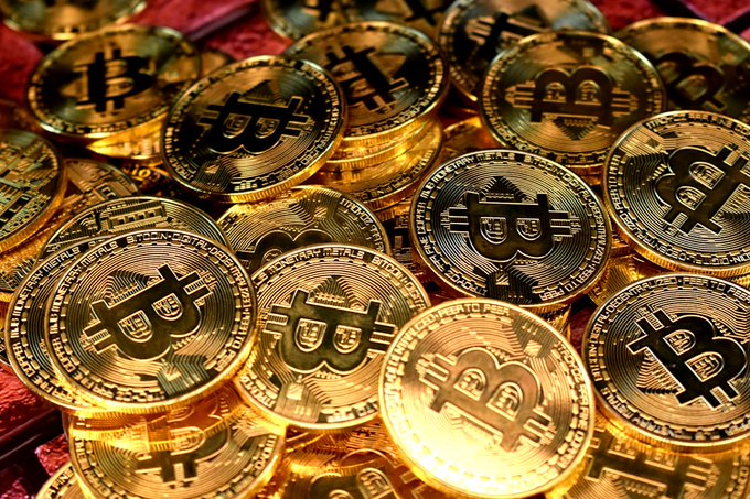 A close-up photograph of a large pile of physical gold-colored Bitcoin tokens stacked together, with the Bitcoin 'B' symbol clearly visible on each coin.