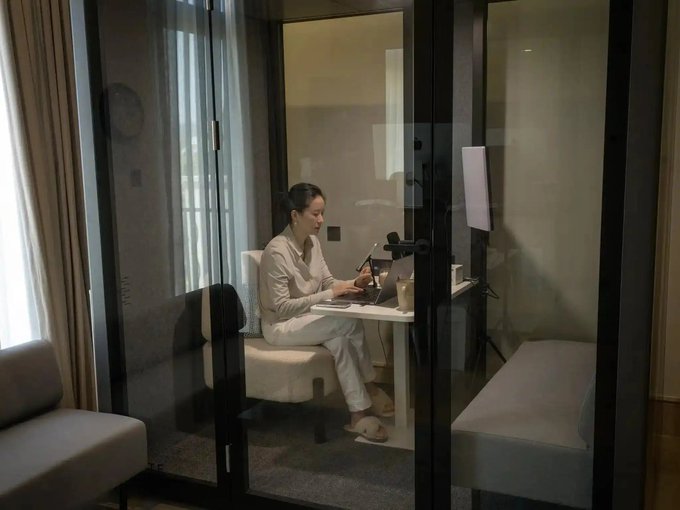 A woman sits in pajamas working on a laptop inside a glass-walled hotel room booth, with a ring light set up for a video call.