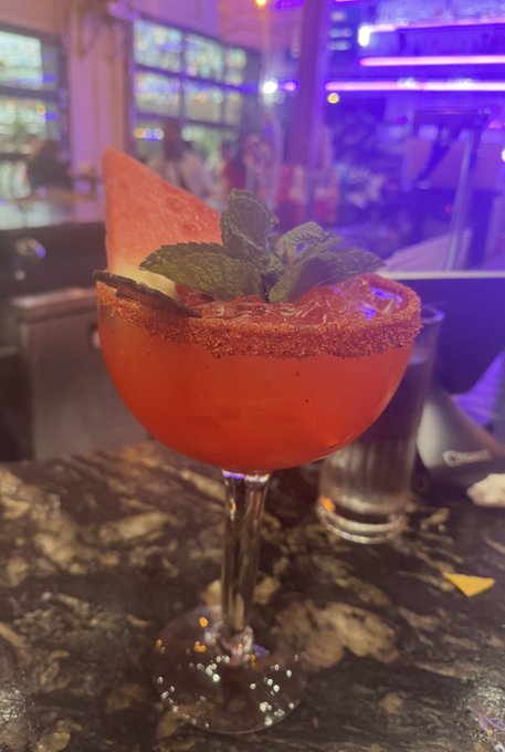 A red cocktail with a spiced rim, garnished with mint, watermelon slice, and citrus, served in a coupe glass on a marble bar top.