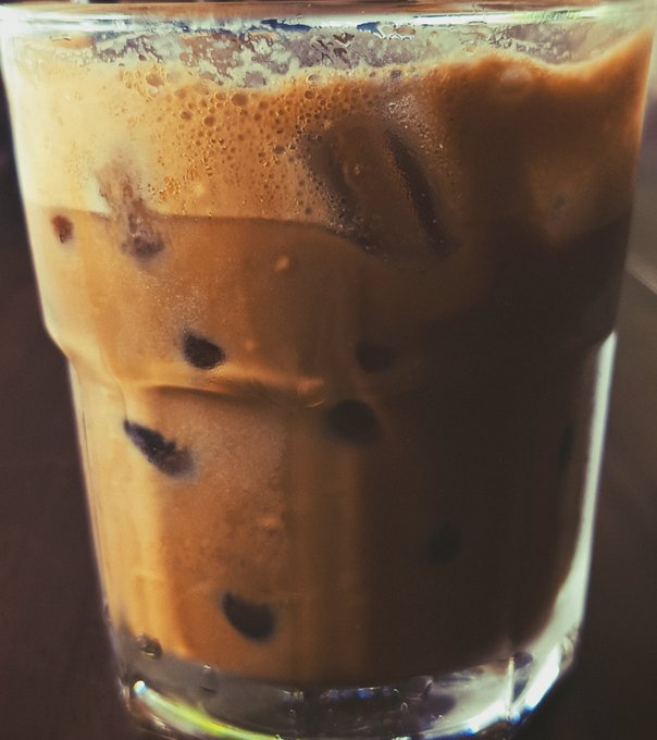 A close-up photograph of a glass filled with iced milk tea or coffee, showing ice cubes and dark tapioca pearls or coffee beans at the bottom.
