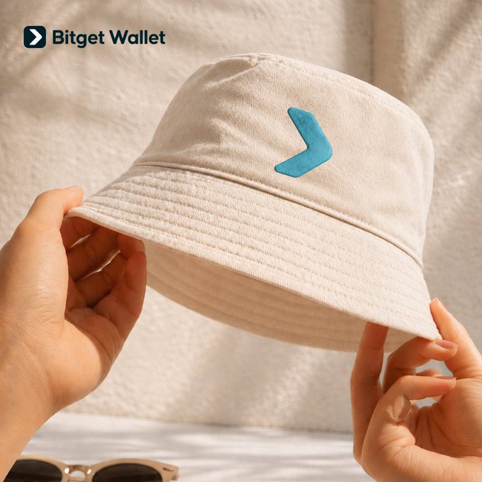 A Bitget Wallet branded cream-colored bucket hat with a turquoise logo, held up by two hands against a soft fabric background.