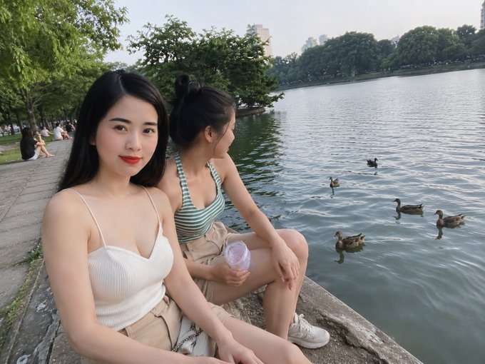 The image features two women sitting by a lake, enjoying each other's company while watching ducks swimming in the water. The women are dressed casually, with one wearing a white tank top and the other wearing a striped shirt. They seem relaxed as they observe the ducks in the pond.