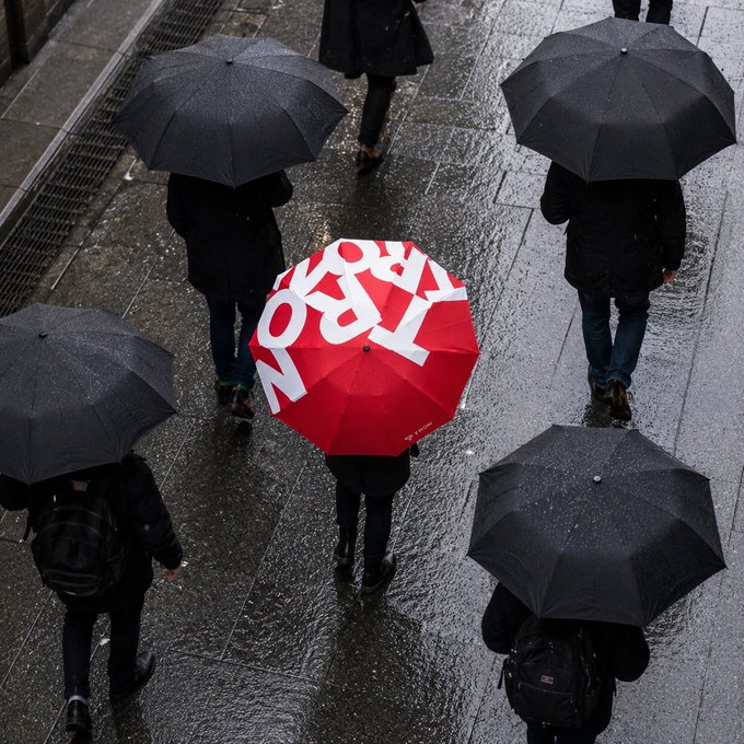 An overhead view of people walking with black umbrellas on a rainy day, with one striking red umbrella in the center displaying white text, creating a visual contrast.