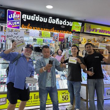 Four people pose cheerfully holding cards in a Thai lottery shop with signs in Thai script including "ศูนย์ซ่อม ป๋อจือฮวน" and price tags like "56".