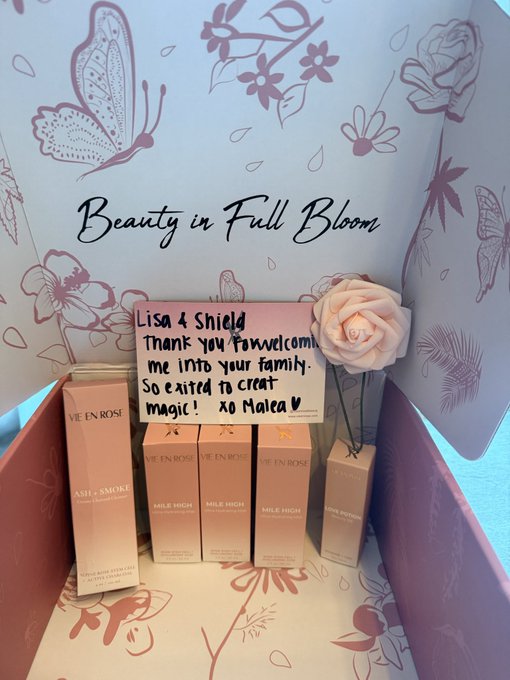 An open Live in Rose 'Beauty in Full Bloom' welcome box containing a hand-written thank you note to Lisa & Shield from Malea, along with sample products including Ash & Smoke and Mile High items, decorated with a pink rose.