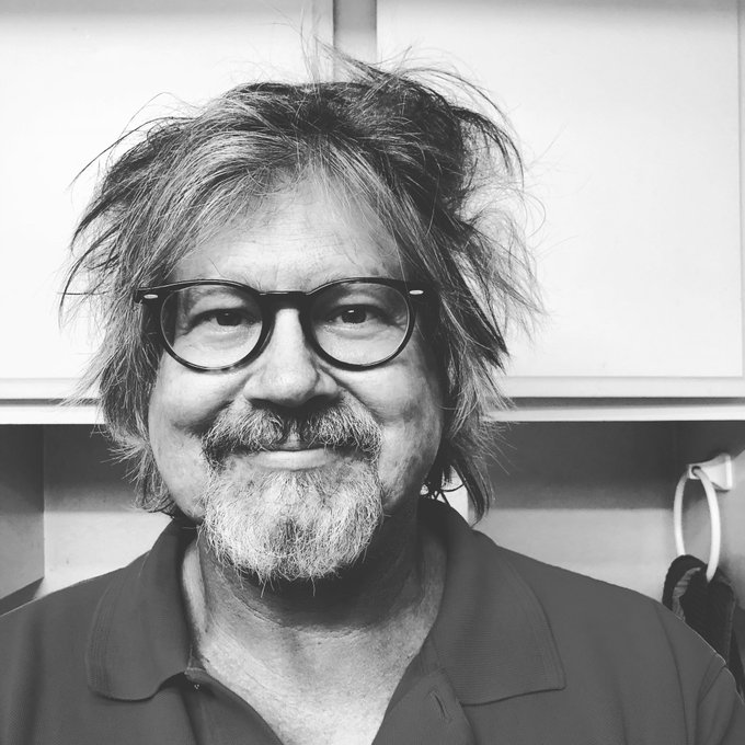 A black-and-white portrait of a middle-aged man with messy hair, a goatee, and round glasses, smiling at the camera.