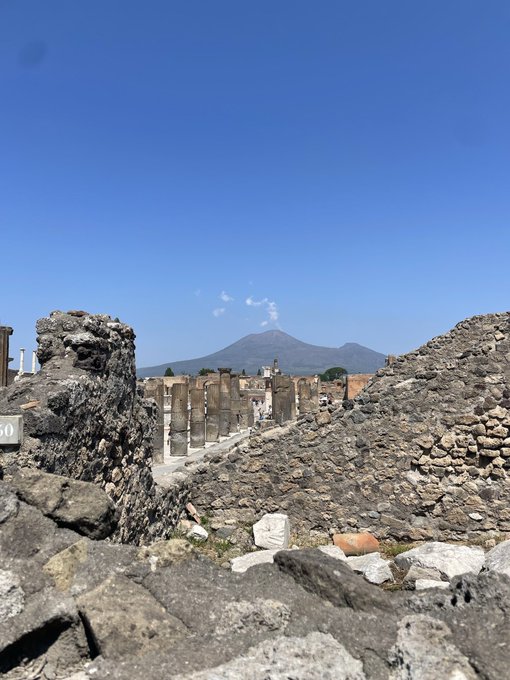 Ancient stone ruins with columns and rubble in the foreground, with a large mountain and clear blue sky in the background, possibly the archaeological site of Pompeii with Mount Vesuvius visible.