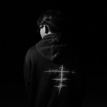 A person in a black hoodie with a white cross design stands with their back to the camera in a dark setting.