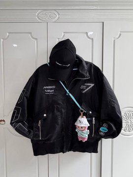 A black hooded jacket with a jagged zipper and logos hangs on a white door, with a small plush keychain attached to it.