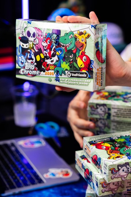 The image features a person holding up a box containing a collection of colorful cartoon characters, likely from the video game "Topps". The box has a vibrant green cover with various cartoon characters on it. The text on the box indicates that this is the global release of "Topps Chrome VeeFriends!". The person's hand is visible in the image, holding up the box against a backdrop of a laptop and other objects.