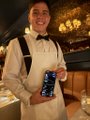 A low-resolution image of a man wearing a white shirt and bow tie holding a blue can, with other people and a picture on the wall in the background.