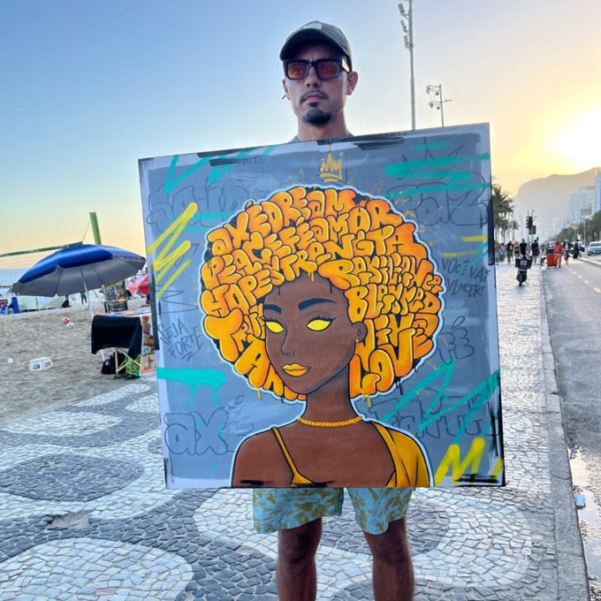 A man wearing sunglasses and a cap stands on a beach boardwalk at sunset, holding up a clear artwork featuring a stylized portrait with an orange afro made of words.