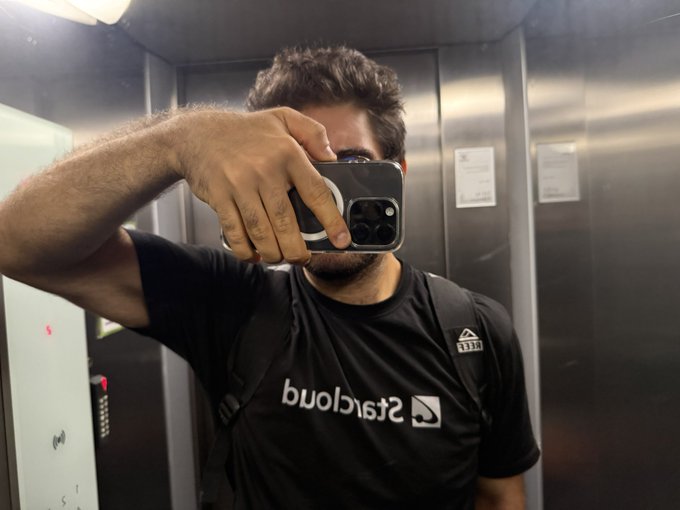 A mirror selfie of a bearded man in an elevator wearing a black 'StayCloud' t-shirt and holding an iPhone with a MagSafe ring.
