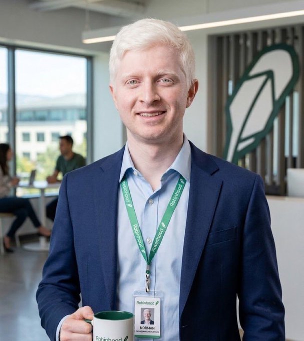A smiling man with white hair, wearing a navy blazer and a Robinhood lanyard with an ID badge labeled "NORMAN," holds a Robinhood-branded mug in a modern office setting, seemingly unaware of the playful threat to "get slimed out on the Robinhood chain" referenced in the caption.