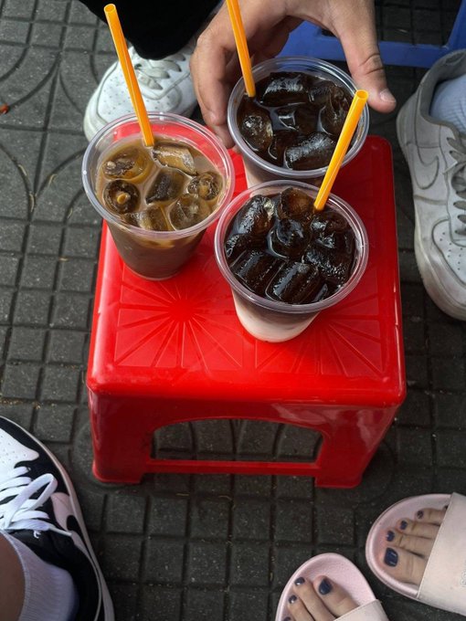 Three clear plastic cups filled with iced coffee drinks and yellow straws sitting on a small red plastic stool, with the feet and sneakers of bystanders visible on the paved ground around it.