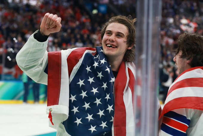 A young man draped in an American flag celebrates with a triumphant fist pump, beaming with joy alongside a teammate, likely after a USA hockey victory at the Olympics. The image captures the patriotic fervor referenced in the post about "americans putting in the $FWA bottom every single day," symbolizing national pride and dedication.