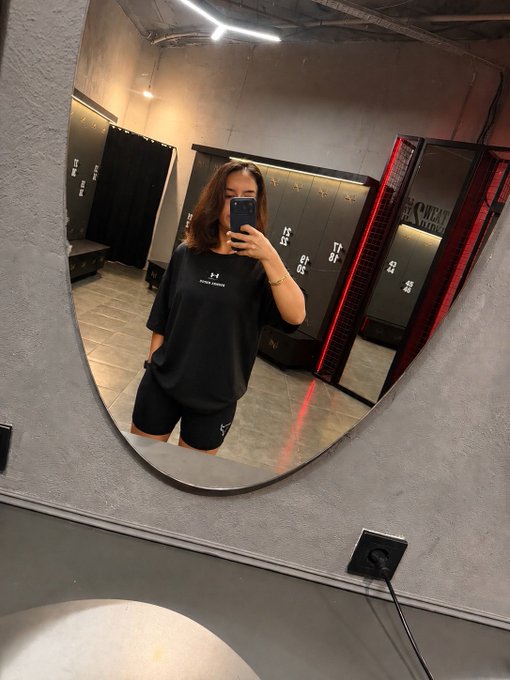A mirror selfie of a woman wearing glasses, a black Under Armour t-shirt, and black shorts, taking the photo with her phone inside what appears to be a locker room.