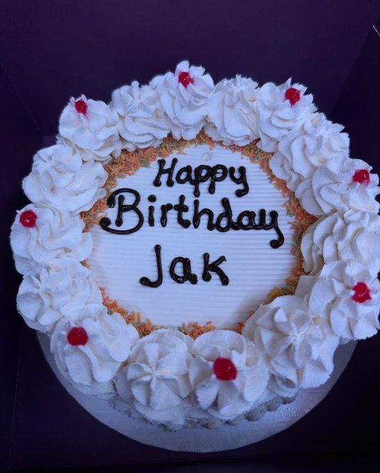 A white birthday cake decorated with frosting rosettes and red cherries, with 'Happy Birthday Jak' written in chocolate icing.
