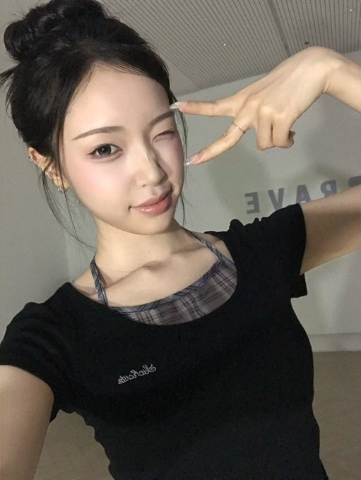 A young woman with her hair in a bun makes a peace sign near her face, wearing a black top with a plaid halter underneath, in a selfie with 'BRAVE' text visible in the background.