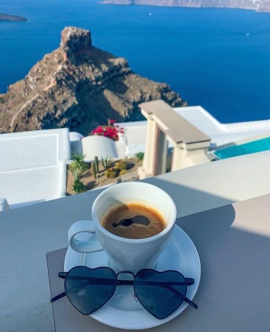 A cup of espresso and heart-shaped sunglasses sit on a white ledge overlooking the stunning blue Aegean Sea and rocky cliffs of Santorini, evoking a serene "good morning" vibe. The tranquil, luxurious scene pairs with the post's optimistic tone about Beldex's growing ecosystem, symbolizing a calm start to a promising day for $BDX.