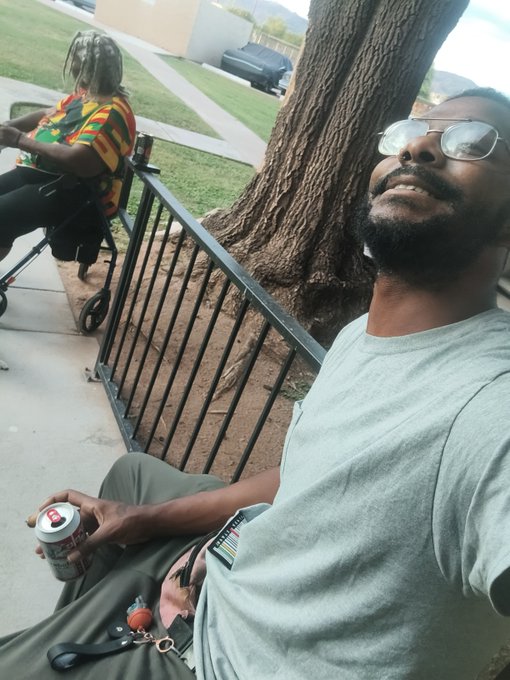 A man wearing glasses and a gray t-shirt sits on a park bench holding a beer can and a book, looking upward, with another person in a colorful shirt seated behind him near a tree.