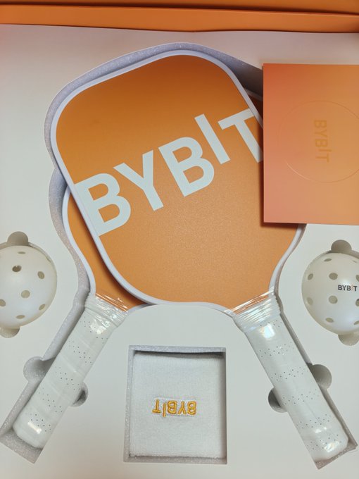 A branded merchandise set featuring two orange and white 'BYBIT' pickleball paddles, white 'BYBIT' pickleballs, and a small 'BYBIT' emblem accessory displayed in a presentation box.