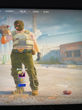 A screenshot from the video game Delta Force shows a player character in tactical gear holding a grenade, standing in an outdoor environment with buildings in the background.