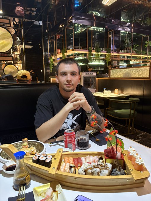 A young man with short dark hair and arm tattoos sits at a restaurant table wearing a black graphic t-shirt, with an elaborate sushi platter served on a wooden boat-shaped platter in front of him, along with a can of Coca-Cola and a glass of dark soda, in a stylish restaurant with mirrored decor and tropical plants.