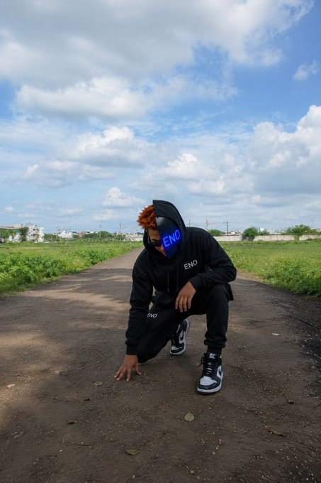 A photo of a person wearing a black hoodie and an 'ENO' mask crouching on an empty road with a cloudy sky background.