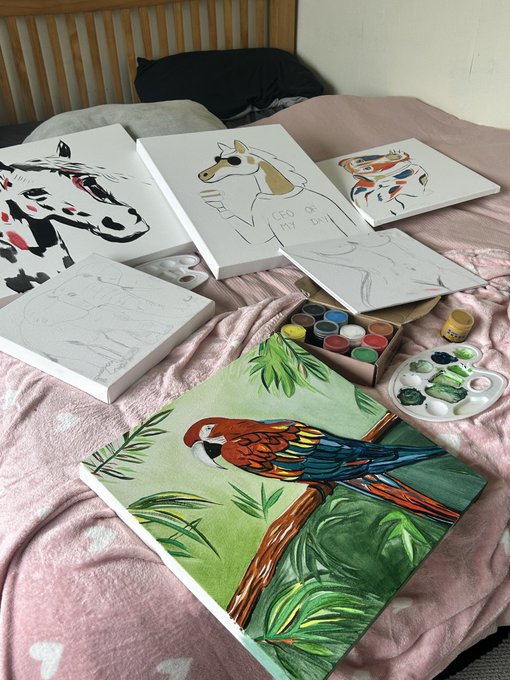 Several canvas paintings rest on a pink bedspread, featuring subjects like a parrot, horse, and shark, alongside a watercolor palette and brushes.