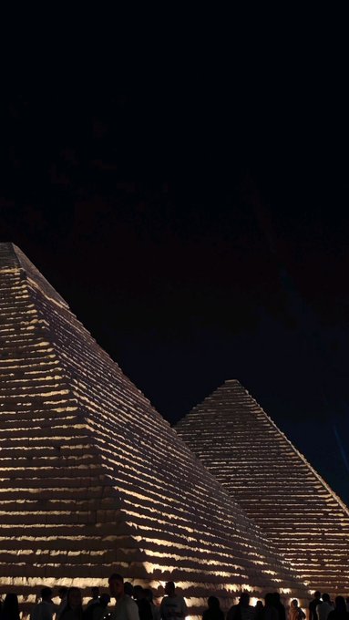 A nighttime photograph of the Pyramids of Giza illuminated with warm, golden light, showing two pyramids and a crowd of small silhouettes at the bottom.