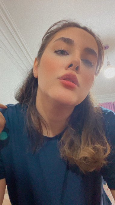 A selfie of a woman with long brown hair, wearing pink lipstick and a dark blue top, posing indoors with a ceiling light visible in the background.
