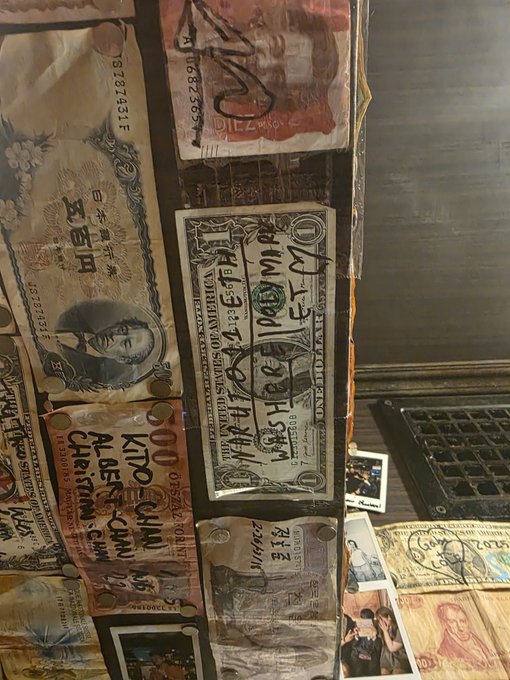 A wall display featuring various international banknotes including Japanese yen and 'KIDOCININ' currency, with a US one-dollar bill in the center covered in handwritten text and signatures.