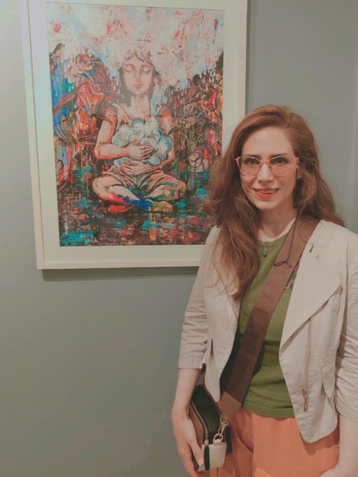 A smiling woman with glasses and a beige jacket poses in an art gallery next to a colorful painting of a seated figure with closed eyes.