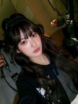 A selfie of an Asian woman with long black hair and bangs, wearing a black jacket, holding a phone in what appears to be a car interior.
