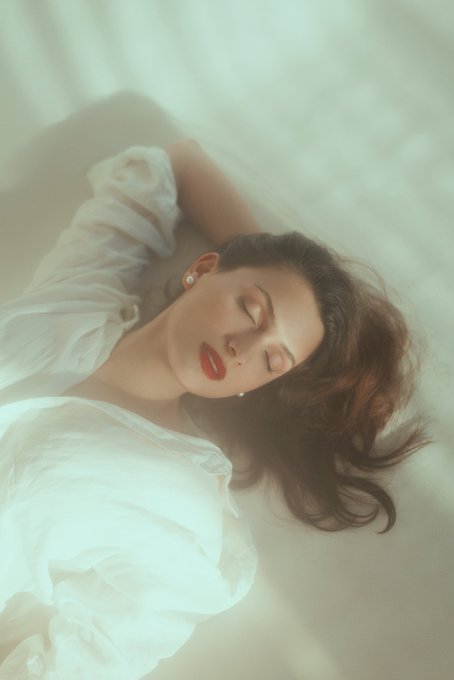 An ethereal portrait of a woman with red lips and closed eyes, wearing a white blouse, floating or resting on a soft cream-colored surface.