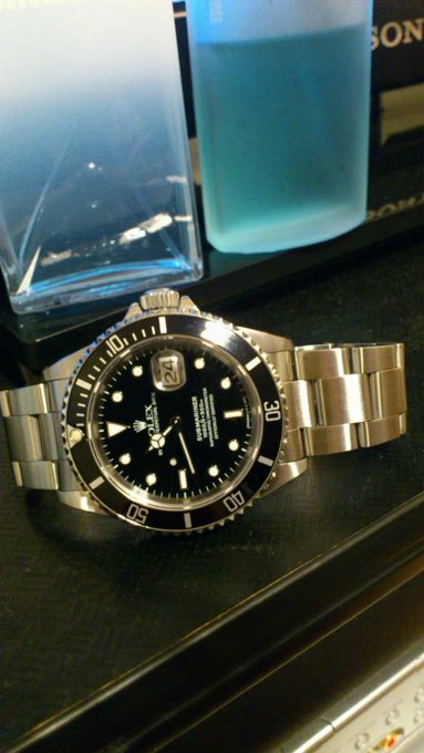 A photograph of a Rolex Submariner watch with a black dial and date display, sitting on a dark surface next to a blue water bottle and what appears to be a glass award.