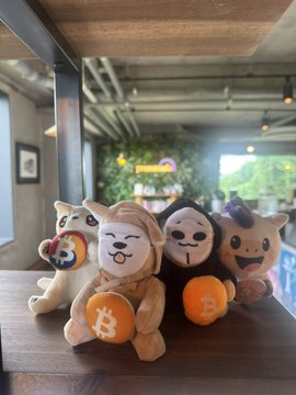 A collection of plush toys including dog figures and a bear figure, displayed on a wooden table with two holding Bitcoin-themed orange coins.