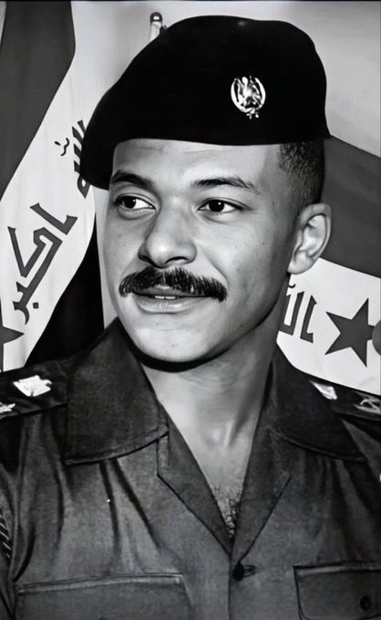 A black and white portrait photograph of a military officer with a mustache wearing a beret and uniform with insignia, standing in front of a flag with Arabic text.