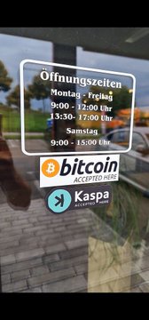 A glass storefront window displaying opening hours in German along with stickers indicating that Bitcoin and Kaspa cryptocurrencies are accepted.