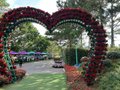 A heart-shaped archway decorated with red flowers framing a pathway leading toward a building, with greenery visible in the background.