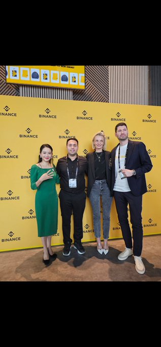 Four people pose in front of a Binance-branded backdrop, including a woman in a green dress holding a smartphone, alongside event attendees wearing lanyards, at what appears to be a Binance industry event. The image aligns with the post's theme of featuring a Binance spokesperson ("@heyibinance") at a corporate gathering, with the prominent Binance logos reinforcing the company's presence and brand visibility at the event.