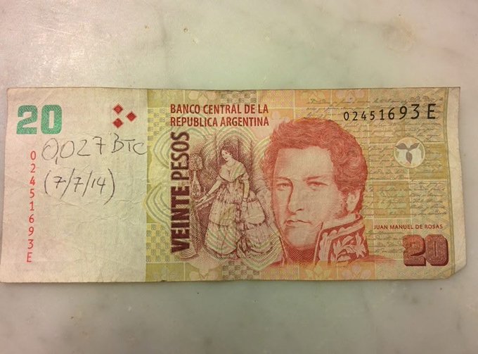 A worn and creased Argentine 20 peso banknote featuring Juan Manuel de Rosas, serial number 02451693 E, has handwritten notations "0027 BTC" and the date "(7/7/14)" on its left side, documenting the historical exchange rate where 20 ARS equaled 0.027 bitcoin.