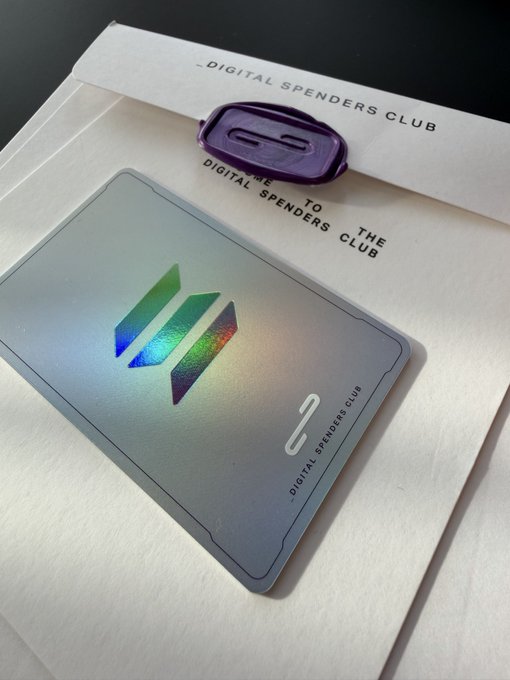 A holographic silver membership card with a colorful logo sits on paper reading 'DIGITAL SPENDERS CLUB' and 'WELCOME TO THE DIGITAL SPENDERS CLUB', with a small purple metallic object nearby.